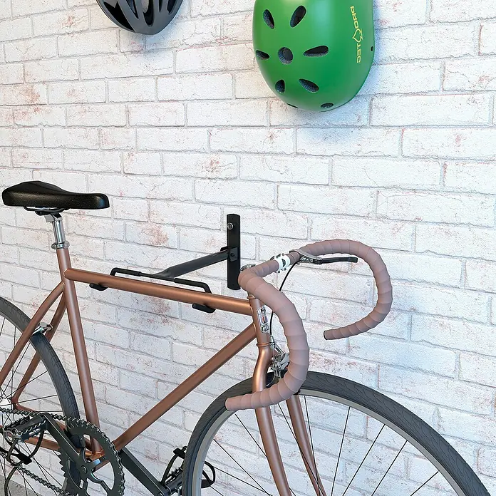 Rei Soporte de pared de bicicletas - L x An x Al: 9 x 32 x 45 cm, Acero, Montaje en la pared - Un portabicicletas montado en una pared de ladrillos, con una bicicleta de carretera colgando, junto con dos cascos encima.