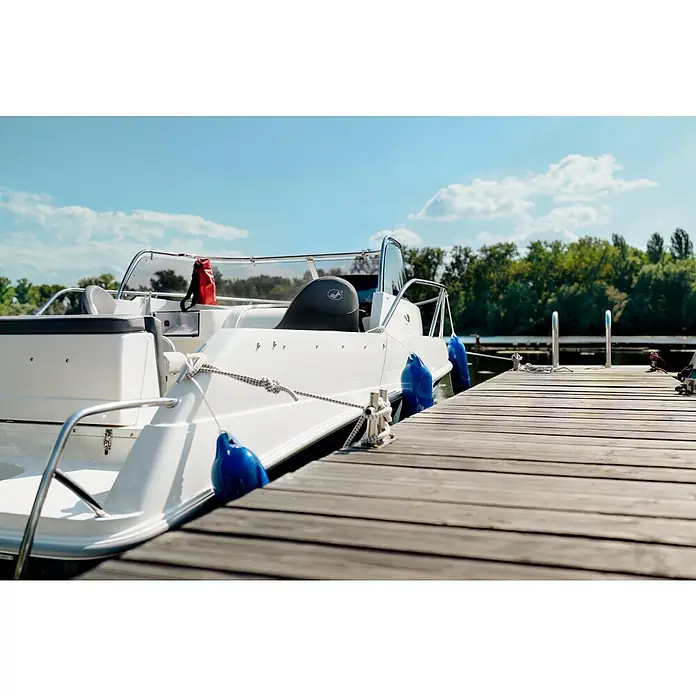 PEGAZUS GFK-Boot 550 Comfort - Umfangreiche Zusatzausstattung, Geeignet für: Max. 6 Personen, Nutzlast: 670 kg, CE-Klasse C - Nahaufnahme eines weißen Motorboots, das an einem Holzsteg mit blauen Fendern und weißen Seilen festgemacht ist, mit Bäumen und blauem Himmel im Hintergrund.