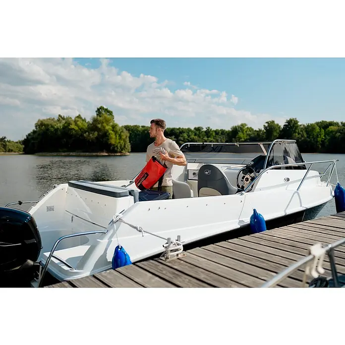 PEGAZUS GFK-Boot 550 Comfort - Umfangreiche Zusatzausstattung, Geeignet für: Max. 6 Personen, Nutzlast: 670 kg, CE-Klasse C - Ein Mann mit einer roten wasserdichten Nautic-Tasche steht auf einem Boot, das an einem Holzsteg vertäut ist. Schönes Wetter und Landschaft.