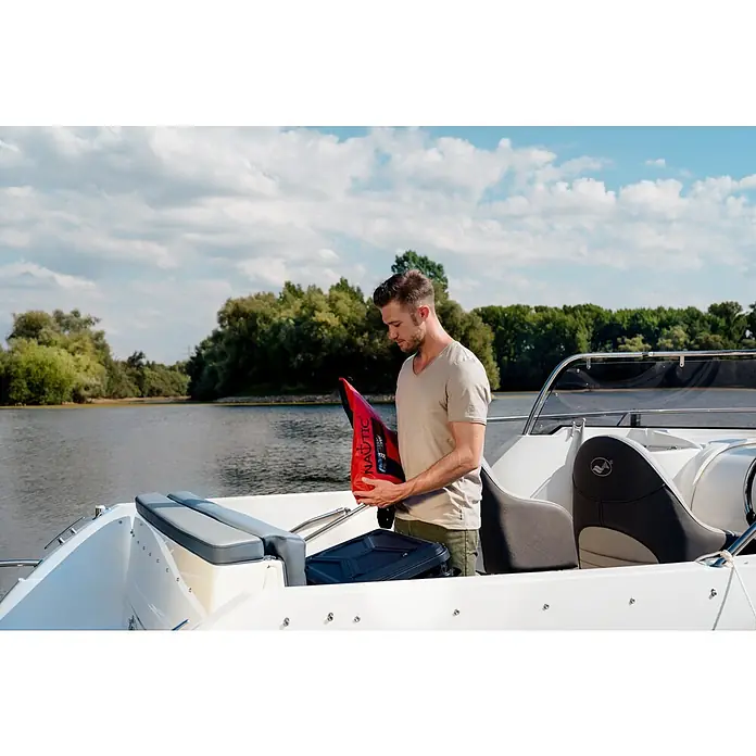 PEGAZUS GFK-Boot 550 Comfort - Umfangreiche Zusatzausstattung, Geeignet für: Max. 6 Personen, Nutzlast: 670 kg, CE-Klasse C - Ein Mann inspiziert eine rote Nautica-Schwimmweste auf einem Boot vor einer malerischen Landschaft mit Bäumen und Wasser.