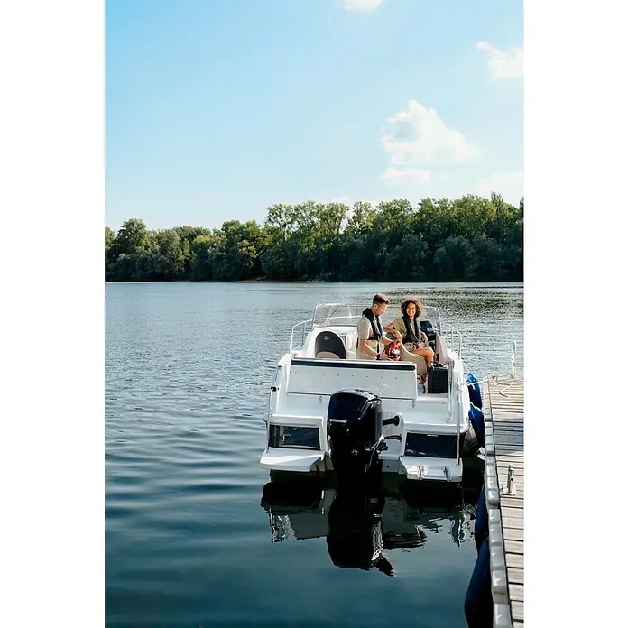 PEGAZUS GFK-Boot 550 Comfort - Umfangreiche Zusatzausstattung, Geeignet für: Max. 6 Personen, Nutzlast: 670 kg, CE-Klasse C - Ein modernes weißes Motorboot liegt an einem Holzsteg mit einem Paar an Bord, vor einer grünen Uferlinie und unter blauem Himmel.