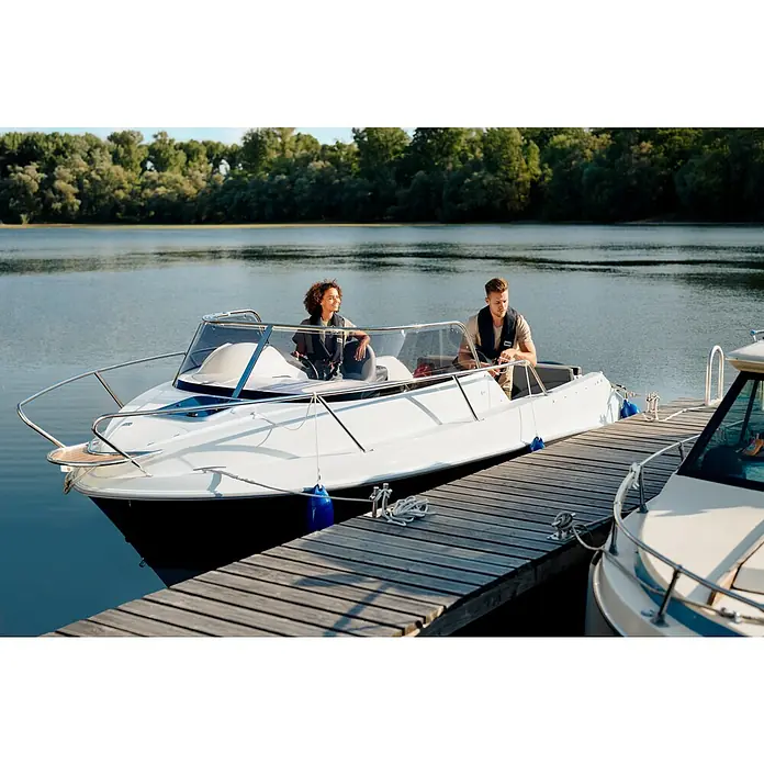 PEGAZUS GFK-Boot 550 Comfort - Umfangreiche Zusatzausstattung, Geeignet für: Max. 6 Personen, Nutzlast: 670 kg, CE-Klasse C - Ein Paar genießt eine Bootsfahrt auf einem weißen Motorboot, das an einem Holzsteg festgemacht ist, mit Bäumen im Hintergrund.