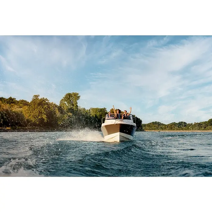 PEGAZUS GFK-Boot 550 Comfort - Umfangreiche Zusatzausstattung, Geeignet für: Max. 6 Personen, Nutzlast: 670 kg, CE-Klasse C - Eine Gruppe von Leuten genießt eine Bootsfahrt auf einem Motorboot, das Wellen auf dem Wasser erzeugt, mit Bäumen im Hintergrund.
