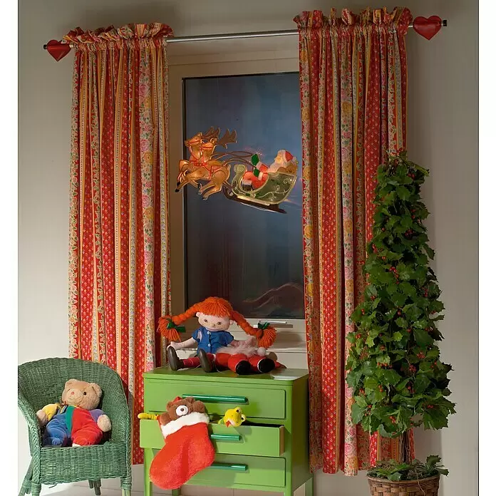 Konstsmide LED-Weihnachtsdekoleuchte Rentier mit Weihnachtsmann und Schlitten - Bunt, Netzbetrieben, LED - Ein gemütlicher Raum, weihnachtlich dekoriert mit roten Blumenvorhängen, einer Santa-Schlitten-Fensterleuchte, Stechpalme und Kinderspielzeug.