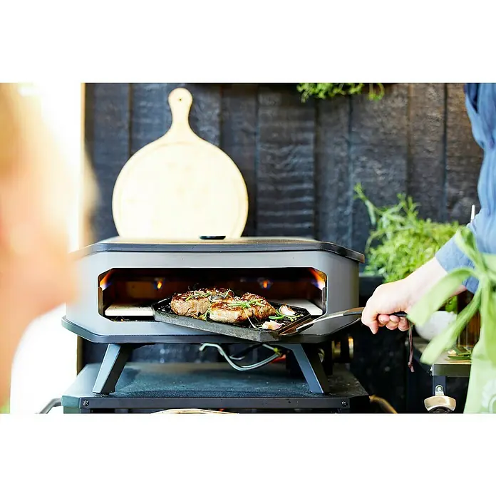 Cozze Horno para pizza gas 17" - 8 kW - Imagen de un horno de pizza exterior en acción, con jugosos filetes cocinándose dentro. El horno está situado contra un fondo de madera rústica.