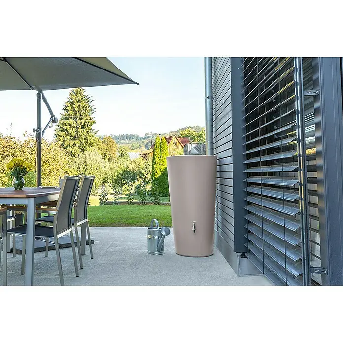 Arves Regenspeicher Rainbowl - 210 l, Taupe - Abgebildet ist ein schlanker, beiger Regenspeicher mit einem Wasserhahn an der Seite, der an einem modernen Haus mit Jalousien steht.