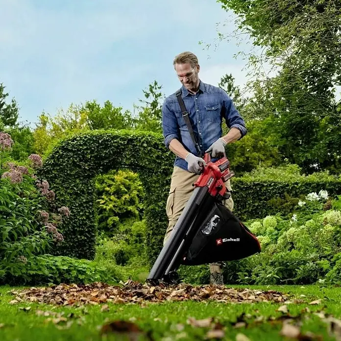 Einhell PXC 18V Soplador/Aspirador de batería Venturro 18/210 - 18 V, Sin batería, Velocidad de soplado: 210 km/h - Un hombre usa un soplador/aspirador de hojas Einhell para limpiar las hojas en el jardín. El dispositivo es rojo y negro.