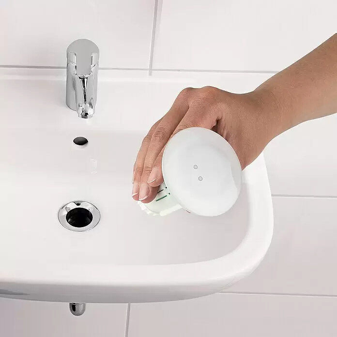 Abfluss-Fee Verschlussstopfen mit LED - Mit Duftstein, Waschbecken - Eine Hand hält ein weißes, rundes Badreinigungsprodukt mit grünem Einsatz über einem weißen Waschbecken.