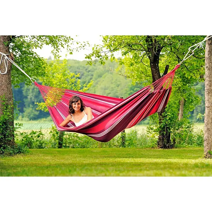 Amazonas Hängematte Salsa fuego - L x B: 310 x 140 cm, Rot/Violett - Frau entspannt in einer rot gestreiften Hängematte, die zwischen zwei Bäumen aufgespannt ist und die idyllische Gartenszene genießt.
