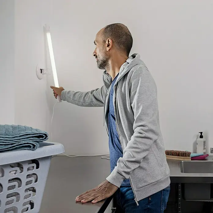 Ledvance LED-Feuchtraumlichtleiste SubmarineLänge: 120 cm, Lichtfarbe: Neutralweiß, 36 W, IP65 - Ein Mann betrachtet eine lange, an der Wand montierte LED-Leuchte. Das Licht scheint hell in einem Waschraum mit Wäschekorb und Spüle.