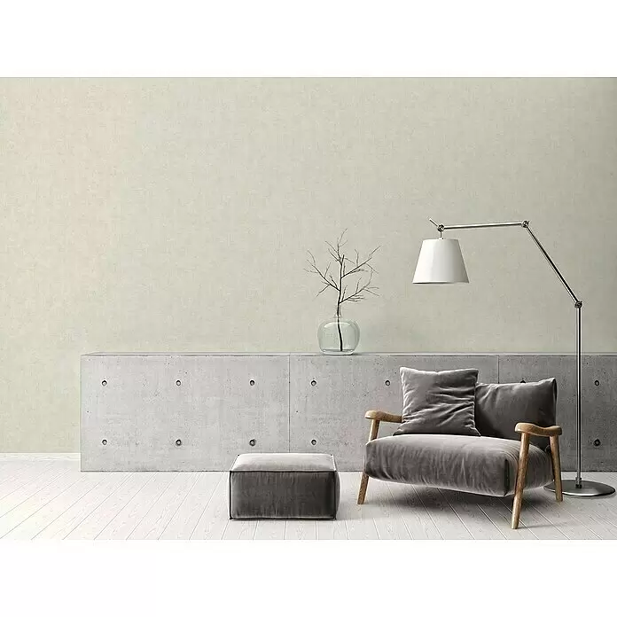A.S. Création Vliestapete - Schattiertes Beige, Uni, 10,05 x 0,53 m - Helle, texturierte Wand in einem modernen Wohnraum mit grauem Sessel, Hocker und Betonkonsole.