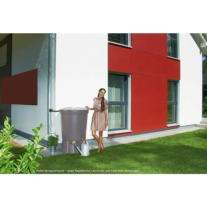 Garantia Basic Line Regentonne Lanzarote - 300 l, Graphit - Eine Frau steht neben einer grauen Regentonne des Modells Lanzarote 300 Liter mit Unterstand vor einem modernen Haus mit roter Fassade.
