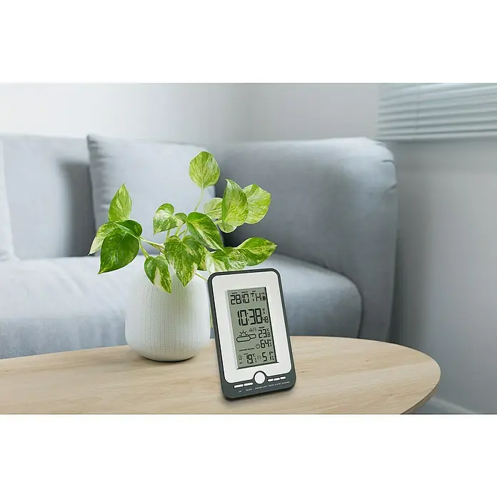 TFA Dostmann Wetterstation Multy - Batteriebetrieben, Grau, Schwarz, 11,8 x 2,9 x 18 cm - Intelligente Wetterstation mit Anzeige von Datum, Uhrzeit, Temperatur und Luftfeuchtigkeit auf einem Tisch mit einer grünen Pflanze.