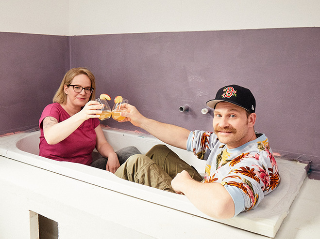 Mann und Frau sitzen bekleidet in Badewanne und stoßen mit einem Getränk an