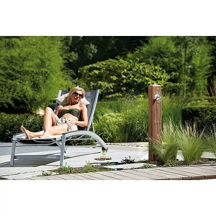 Garantia Wasserzapfstelle Wood - Darkwood, Höhe: 100 cm - Frau entspannt auf einer grauen Metallliege mit hellem Kissen im Garten. Eine Außenzapfsäule ist in der Nähe.