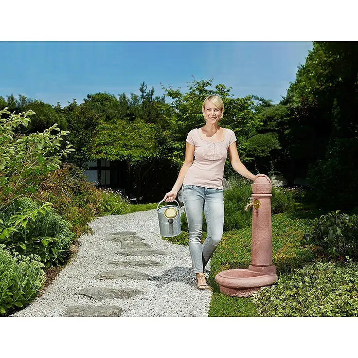 Garantia Brunnen Venezia - Redstone, 52 x 44 x 90 cm - Eine Frau steht in einem Garten mit einem Metalleimer und einer dekorativen Gartenwasserstelle aus Terrakotta.