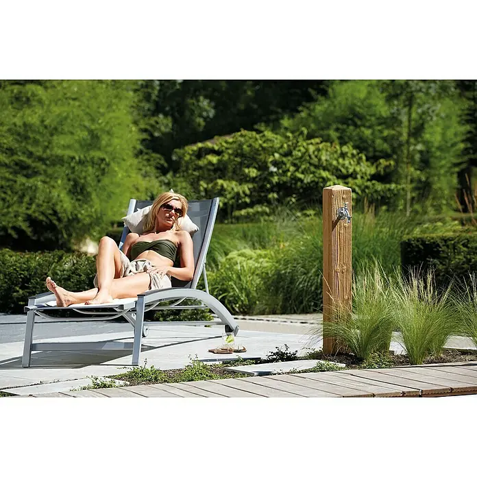 Garantia Wasserzapfstelle Wood - Lightwood, Höhe: 100 cm - Eine Frau entspannt sich auf einer grauen Sonnenliege im Freien, neben einem rustikalen Wasserhahn und üppigem Grün.