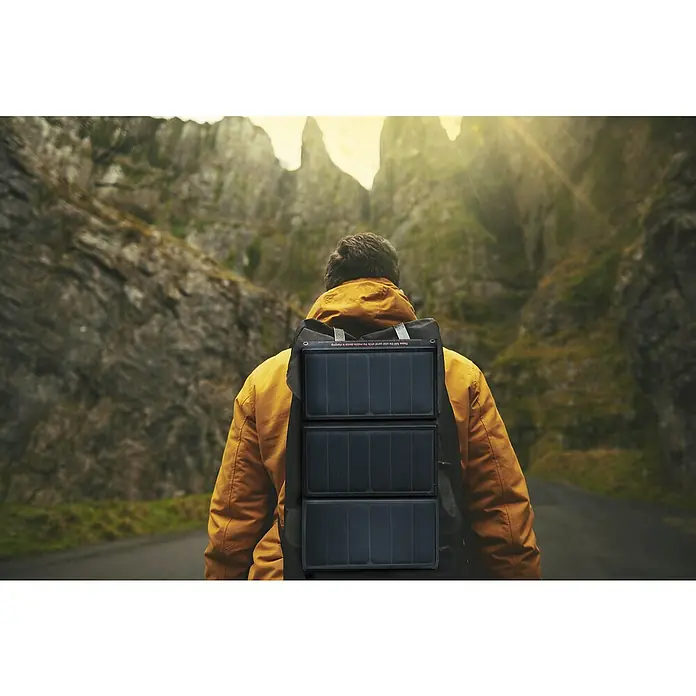Yard Force Solar-Powerbank LX PB21 - 48,1 Wh, 65 x 28,5 x 1,5 cm - Ein Mann mit einem gelben Rucksack mit einem Solarpanel darauf wandert auf einer Straße zwischen Hügeln. Das Solarpanel lädt mobile Geräte auf.