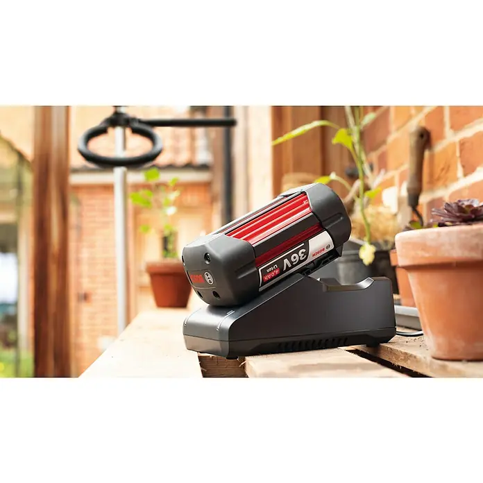 Bosch Power for All 36V Batterie-Ladegerät AL 36V-20 - 36 V, Passend für: alle 36V POWER FOR ALL-Akkus - Ein Bosch 36V Li-Ion Akku befindet sich zum Aufladen auf einem schwarzen Ladegerät im Freien, umgeben von Pflanzen und Gartengeräten.