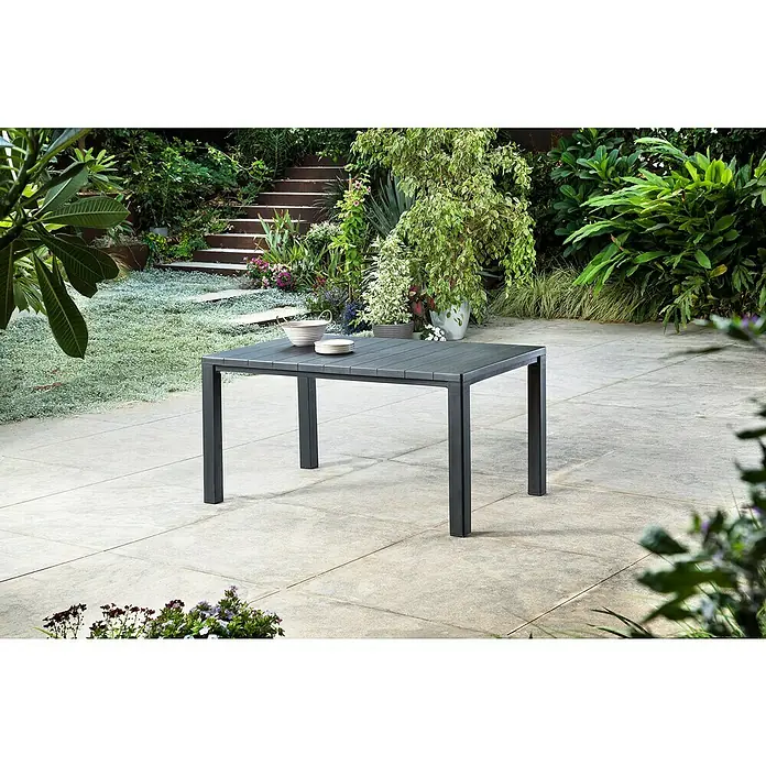 Keter Mesa de jardín Julie - L x An x Al: 149 x 92 x 75 cm, Polipropileno, Grafito - Una mesa de comedor gris para exteriores se encuentra en un patio, rodeada de una exuberante vegetación. Cuencos y platos están sobre la mesa.