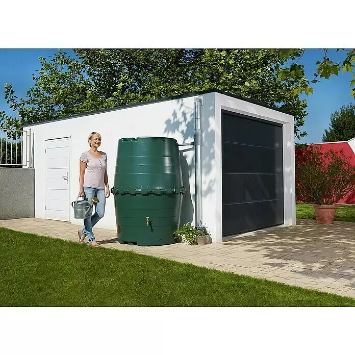 Garantia Basic Line Regentonne - Grün - Eine Frau hält eine Gießkanne neben einer großen grünen Regentonne, die Wasser aus einem Fallrohr sammelt, vor einer modernen Garage.