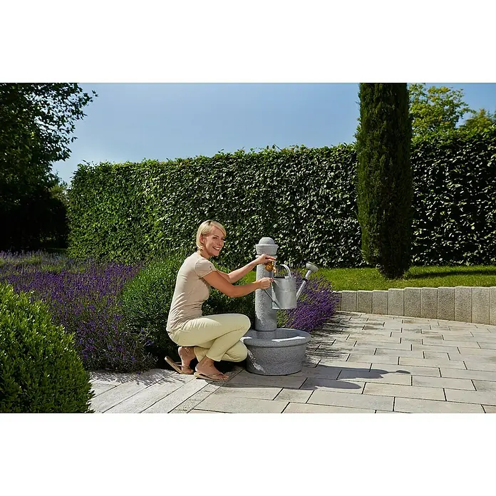 Garantia Brunnen Roma - Lightgranite, 54 x 54 x 100 cm - Frau befüllt eine graue Gießkanne am stilvollen Gartenbrunnen, umgeben von Lavendel und Grünpflanzen.