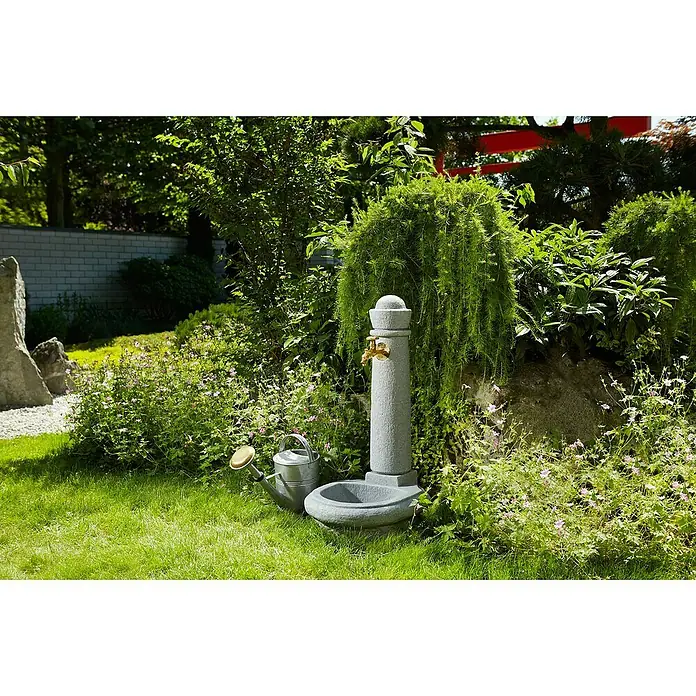 Garantia Brunnen Venezia - Lightgranite, 52 x 44 x 90 cm - Eine Steingarten-Wasserstelle mit messingfarbenem Hahn, umgeben von üppigem Grün und einem nostalgischen Zinkgießkanne.