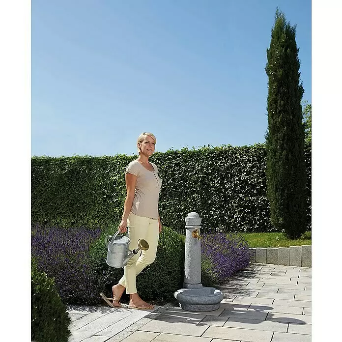 Garantia Brunnen Venezia - Lightgranite, 52 x 44 x 90 cm - Grauer Steingartenbrunnen mit Messinghahn, auf einem gepflasterten Weg in einem Garten mit Lavendel. Eine Frau steht mit einer Gießkanne daneben.