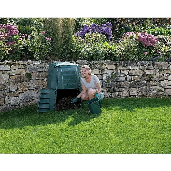 Garantia Komposter Thermo Star - 1.000 l, 130 x 130 x 102 cm - Eine Frau beugt sich zu einem grünen Komposter in einem Garten. Der Komposter steht vor einer Steinmauer und ist mit Grünzeug bedeckt.