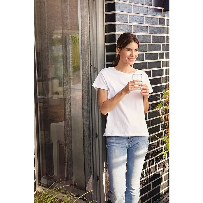 Tesa Insect Stop Lamellenvorhang Premium - B x H: 95 x 220 cm - Frau mit weißem T-Shirt und Jeans, die ein Glas Wasser hält. Helle, freundliche Atmosphäre auf einer Terrasse mit Blick auf eine Tür.