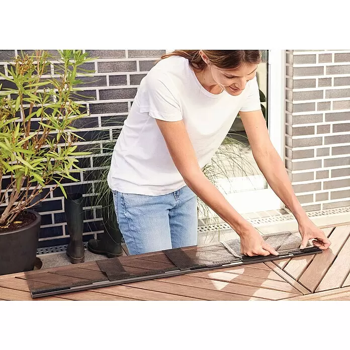 Tesa Insect Stop Lamellenvorhang Premium - B x H: 95 x 220 cm - Eine Frau montiert einen dunkelgrauen, modularen Insektenschutz mit Gitterelementen auf einem Holztisch im Freien.