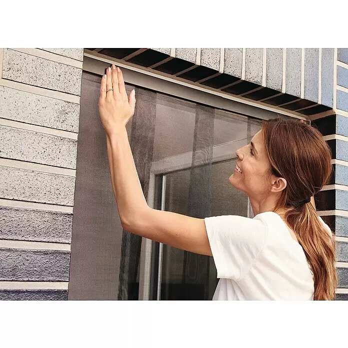 Tesa Insect Stop Lamellenvorhang Premium - B x H: 95 x 220 cm - Eine Frau berührt ein dunkelgraues Insektenschutzgitter aus Mesh, das in einem Fenster installiert ist, und prüft lächelnd dessen Passform.