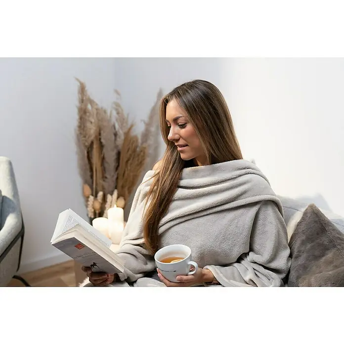 Decke Giny Ärmel - Pumice Stone, 200 x 150 cm, 100 % Polyester - Frau liest ein Buch, eingewickelt in eine weiche, graue Kuscheldecke mit Ärmeln, während sie eine Tasse Tee hält, umgeben von Kerzen und dekorativem Gras.