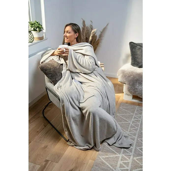 Decke Giny Ärmel - Pumice Stone, 200 x 150 cm, 100 % Polyester - Frau entspannt in einem Sessel mit einer grauen Ärmeldecke und einer Tasse in der Hand, während sie aus dem Fenster schaut.