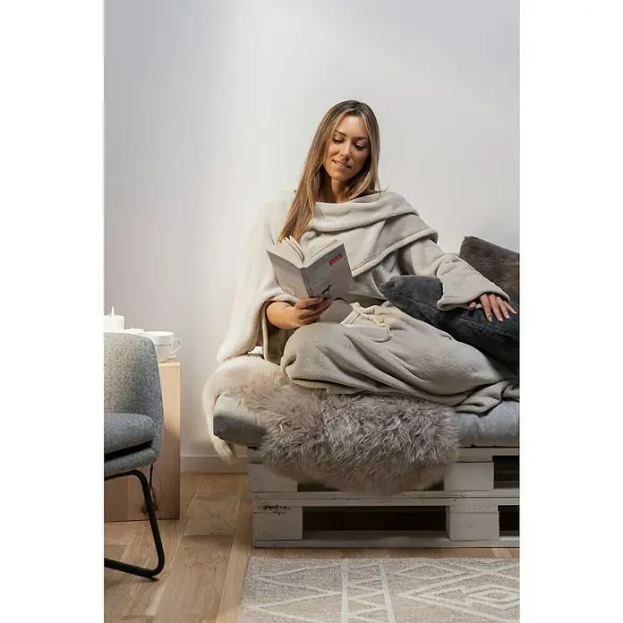 Decke Giny Ärmel - Pumice Stone, 200 x 150 cm, 100 % Polyester - Eine Frau liest in einen beigen Ärmel-Decke gehüllt ein Buch. Die Ärmel-Decke ist kuschelig und hält warm beim Lesen.
