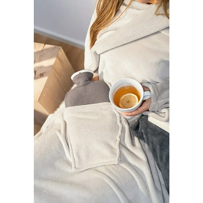 Decke Giny Ärmel - Pumice Stone, 200 x 150 cm, 100 % Polyester - Das Foto zeigt eine Frau mit einer Wärmflasche und einer Tasse Tee. Die Wärmflasche steckt in einer Tasche, die anscheinend an einer Decke befestigt ist.