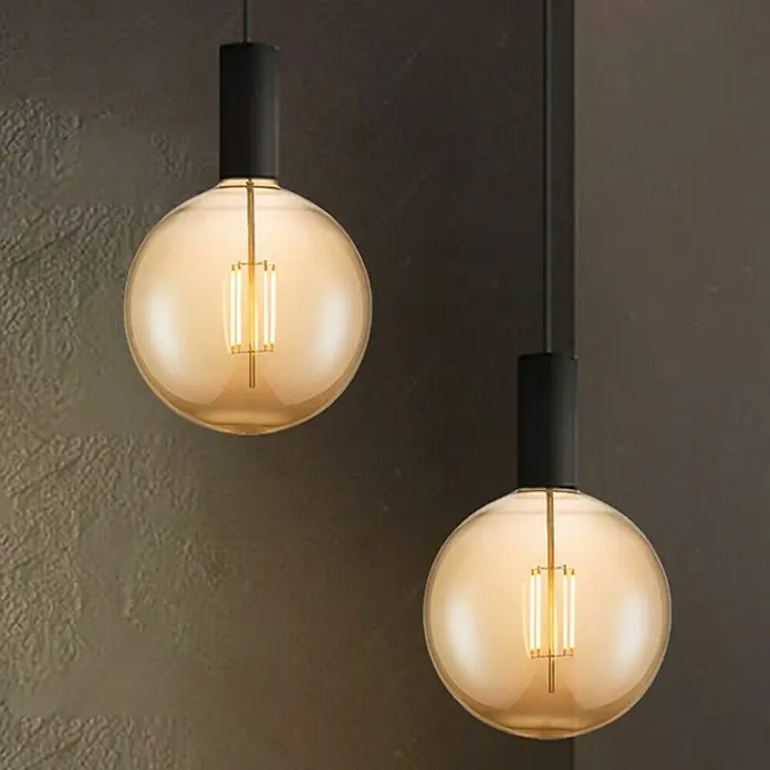 Home Sweet Home LED-Lampe Globe-Form E27 - E27, Dimmbar, Warmweiß, 400 lm, 4 W, Durchmesser: 18 cm - Zwei moderne Pendelleuchten mit bernsteinfarbenen Glaskugeln und schwarzen Aufhängungen vor einer dunklen Wand.