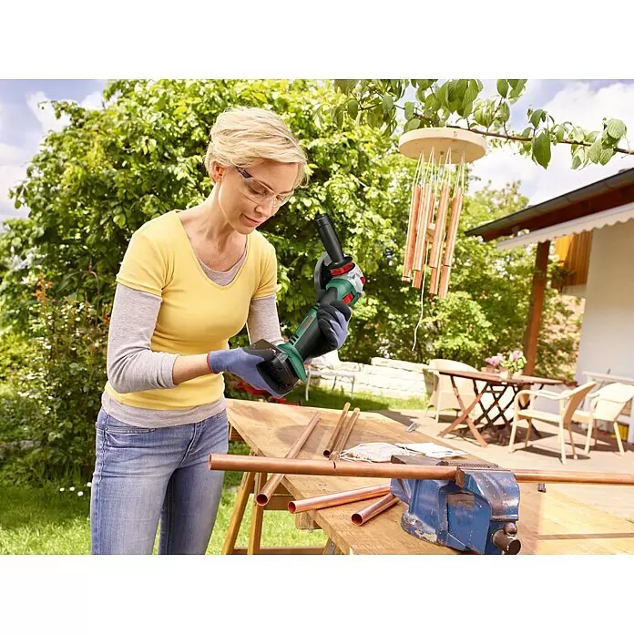 Bosch Power for All 18V Akku-Winkelschleifer AdvancedGrind 18V-80 - 18 V, Ohne Akku, 125 mm, 2.800 U/min - 12.000 U/min - Frau nutzt grüne und schwarze Bosch Winkelschleifer, um Kupferrohre zu schneiden, trägt Schutzbrille und Handschuhe im Freien.