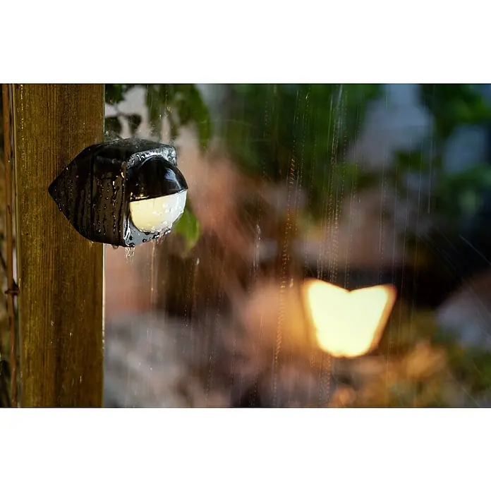 Philips Hue Bewegungsmelder - Reichweite Sensor: 12 m, Schwarz, L x B x H: 7,5 x 17,6 x 16,5 cm - Wasserdichter LED-Gartenstrahler an einem Holzpfosten im Regen, der eine gemütliche und sichere Beleuchtung für den Außenbereich bietet.