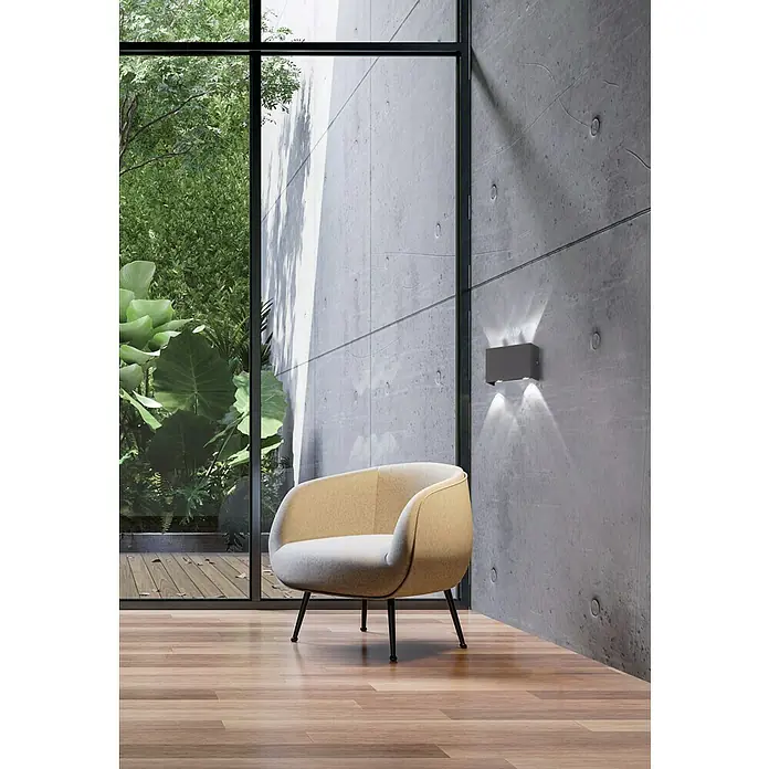 Fischer & Honsel Wandleuchte Nairobi - 10 W, L x B x H: 140 x 24 x 24 cm, Schwarz - Ein moderner Sessel mit hellbeigem Bezug steht auf einem Holzboden vor einer Betonwand mit einer Wandleuchte.
