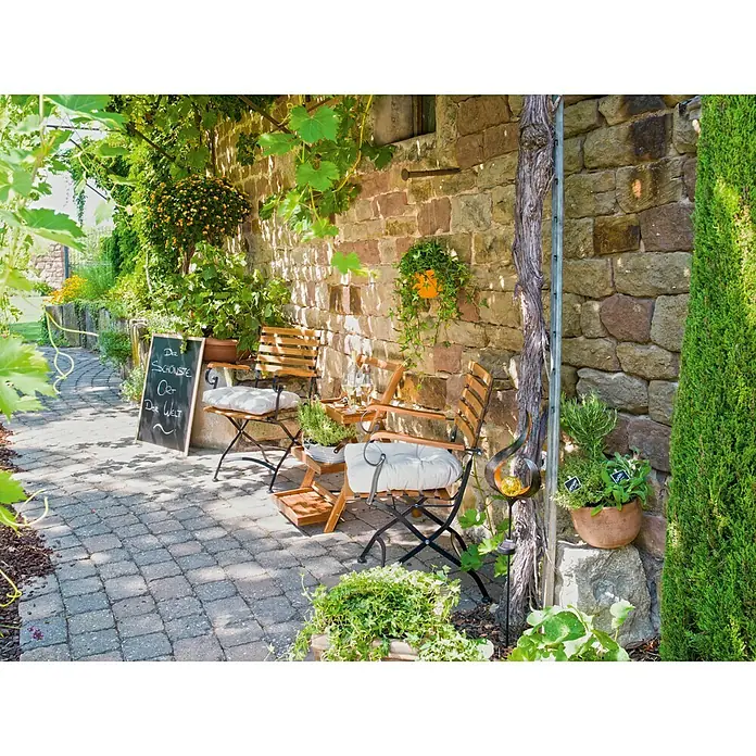 Sunfun Diana Blumentreppe - 40 x 65 x 80 cm, Akazie, Naturbraun - Gemütlicher Gartensitzplatz mit Holzstühlen und Tischchen vor einer Steinmauer mit Pflanzen. Die Outdoor-Möbel laden zum Verweilen ein.