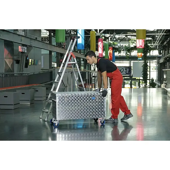 Alutec München Aufbewahrungs- & Transportbox R250 - 1.022 x 525 x 520 mm, 250 l - Aluminium Werkzeugkiste mit Rollen und eine Leiter. Der Arbeiter in roter Arbeitskleidung schiebt die Werkzeugkiste in einer Halle.
