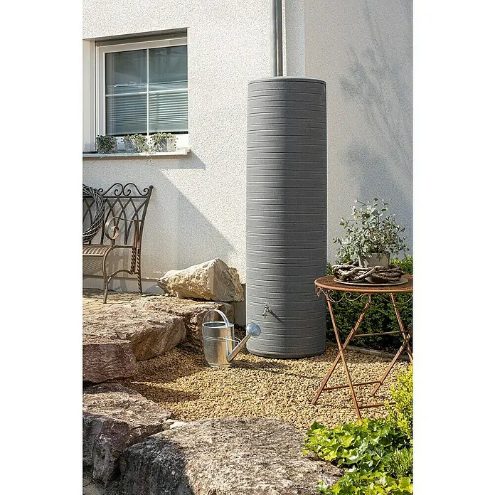 Arves Regenspeicher Novara - 500 l, Black Granit - Grauer Wasserbehälter mit horizontalen Rillen neben einem Haus mit Steinen, einer Gießkanne und einem Beistelltisch.
