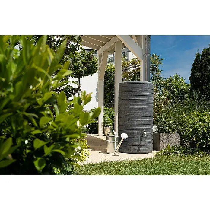 Arves Regenspeicher Novara - 280 l, Black Granit - Nahaufnahme eines Regenspeichers neben einer Gießkanne im Garten. Der Tank ist grau und zylindrisch mit horizontalen Rillen.