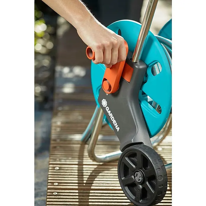 Gardena Carro portamangueras Cleveroll S - Plástico/metal, Capacidad de manguera: 40 m, 13 mm (½″) - Primer plano de una persona operando un enrollador de manguera GARDENA con rueda sobre una terraza de madera.