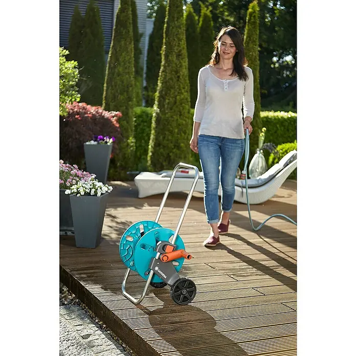 Gardena Carro portamangueras Cleveroll S - Plástico/metal, Capacidad de manguera: 40 m, 13 mm (½″) - Una mujer tira de un carro portamangueras Gardena en una terraza de madera. El carro es azul con detalles grises y un conector naranja.