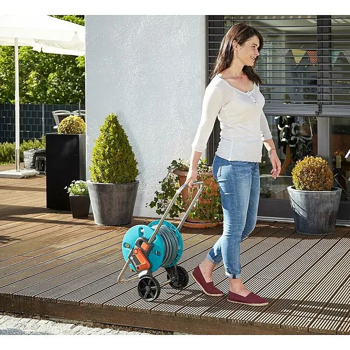 Gardena Carro portamangueras Cleveroll S - Plástico/metal, Capacidad de manguera: 40 m, 13 mm (½″) - Una mujer tira de un carro portamangueras GARDENA por su jardín. El carro es azul y tiene un mango naranja.