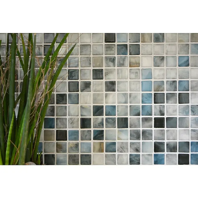 Mosaikfliese Quadrat Eco Air Force VP56256PUR - 31,6 x 31,6 cm, Mix Grau, Glänzend - Nahaufnahme einer Wand mit Mosaikfliesen in verschiedenen Grautönen und Blautönen, ergänzt durch eine grüne Pflanze im Vordergrund.