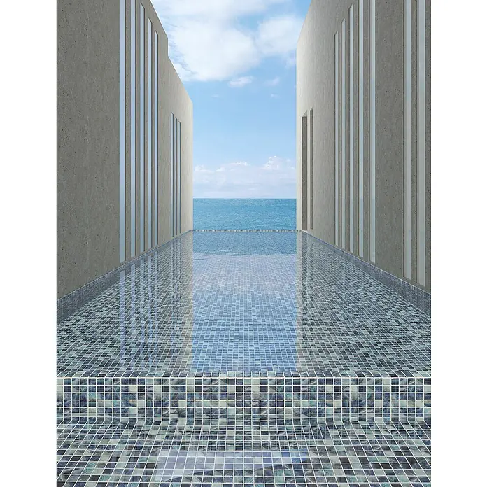 Mosaikfliese Quadrat Eco Royal VP56254PUR - 31,6 x 31,6 cm, Mix Royalblau, Glänzend - Perspektive eines mosaikgefliesten Pools, der sich zum Meer hin erstreckt, flankiert von modernen Gebäuden. Die blauen Mosaikfliesen schaffen einen harmonischen Übergang.