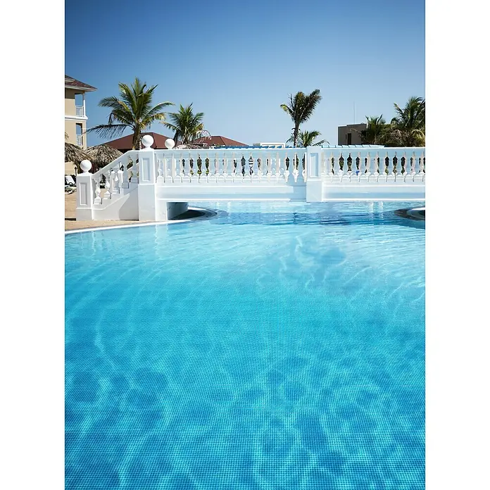 Mosaikfliese Quadrat Crystal Uni GM A 31P - 32,7 x 30,5 cm, Blau, Glänzend - Blick auf ein Schwimmbad mit türkisfarbenem Wasser und einer weißen Brücke. Palmen und Gebäude sind im Hintergrund zu sehen.